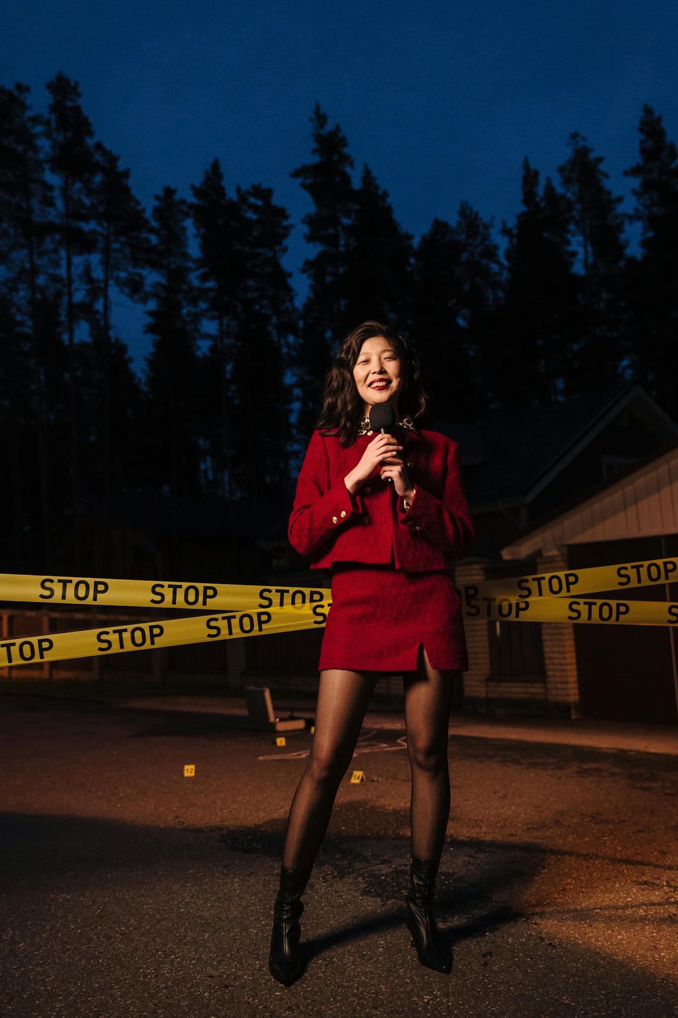 Asian female reporter in red dress reporting with microphone by barricade tape during night.